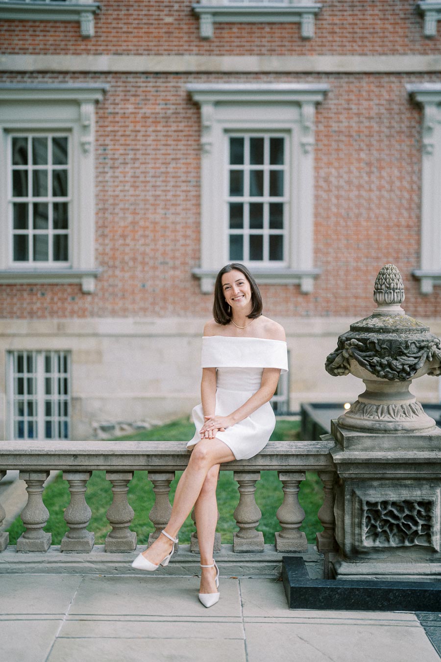 A smiling woman in a white off-the-shoulder dress sitting on a stone balustrade in front of a historic brick building.