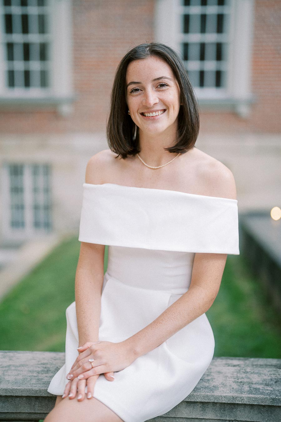 Smiling woman in an elegant off-shoulder white dress sitting outdoors in front of a brick building, showcasing a natural and graceful pose.