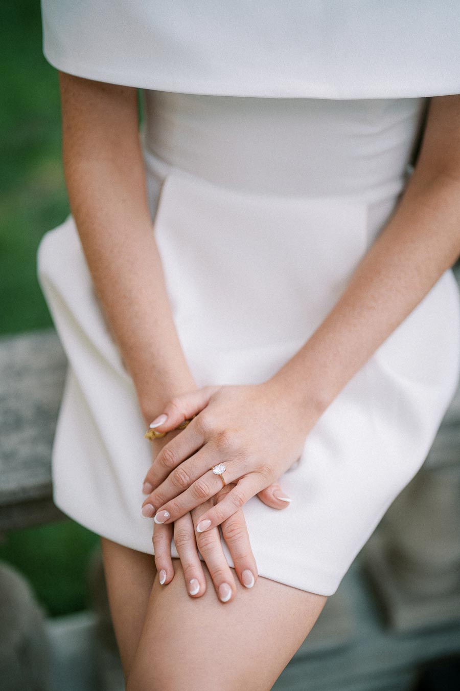 Elegant woman in a white dress with manicured nails, displaying a delicate engagement ring on a sunny day.