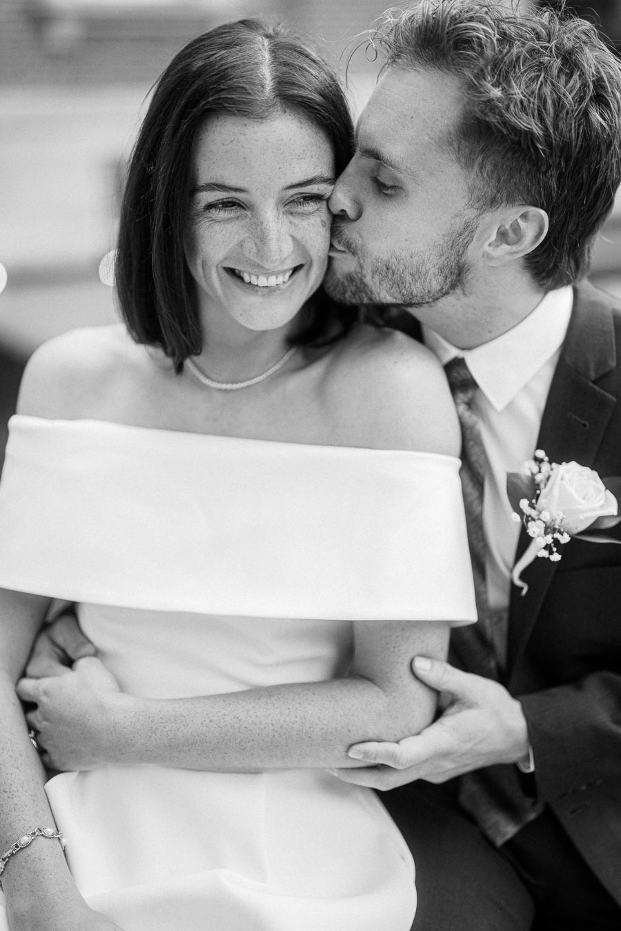 A black and white photo of a smiling bride wearing an off-the-shoulder dress, embraced by her partner in a suit with a floral boutonniere, sharing a tender moment on their wedding day.