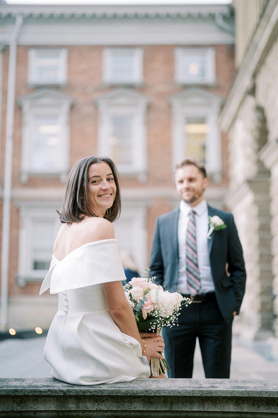 A bride in a white dress smiling while holding a bouquet of flowers, with a man in a suit and tie in the background outside a historic building.