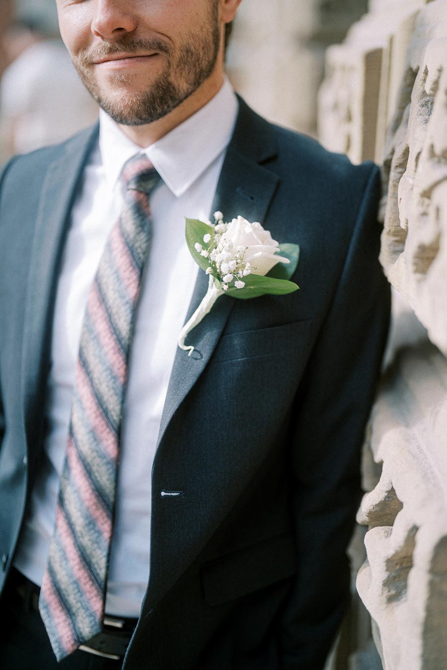 Groom wearing a dark suit and patterned tie with a white rose boutonniere, smiling and leaning against a stone wall.