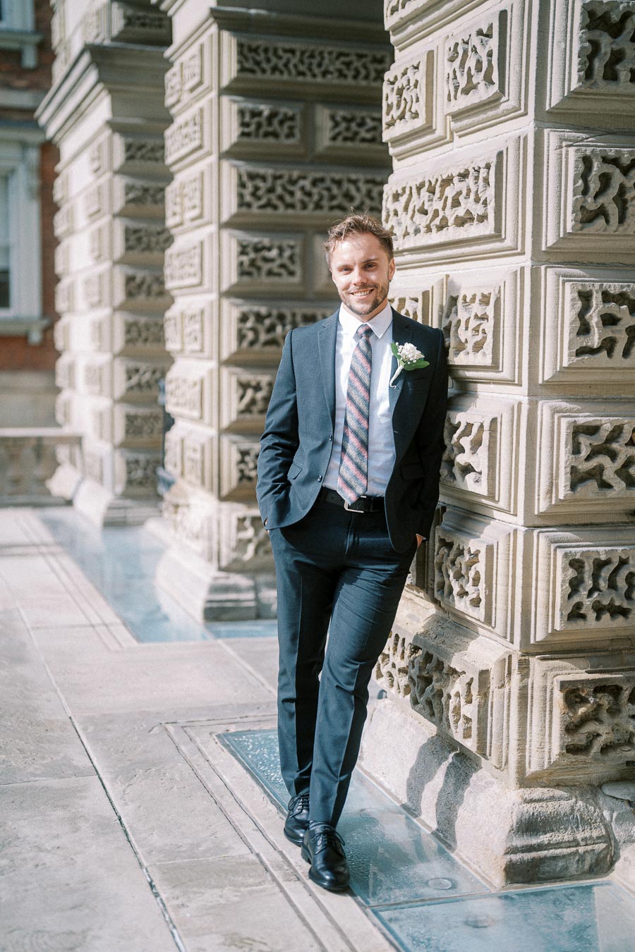 A man in a navy suit with a floral boutonniere stands smiling against a decorative stone pillar of a historic building.