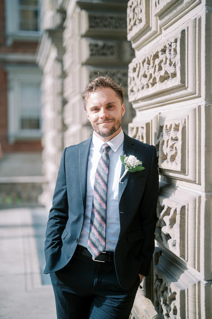 Young man in a suit with a floral boutonniere, standing against an ornate stone wall, smiling confidently at the camera in a bright, outdoor setting.