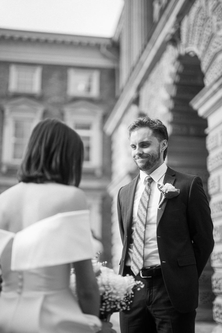 Black and white wedding photo of a bride and groom exchanging vows outdoors, with the groom smiling and wearing a suit and boutonniere, while the bride holds a bouquet in an elegant dress.