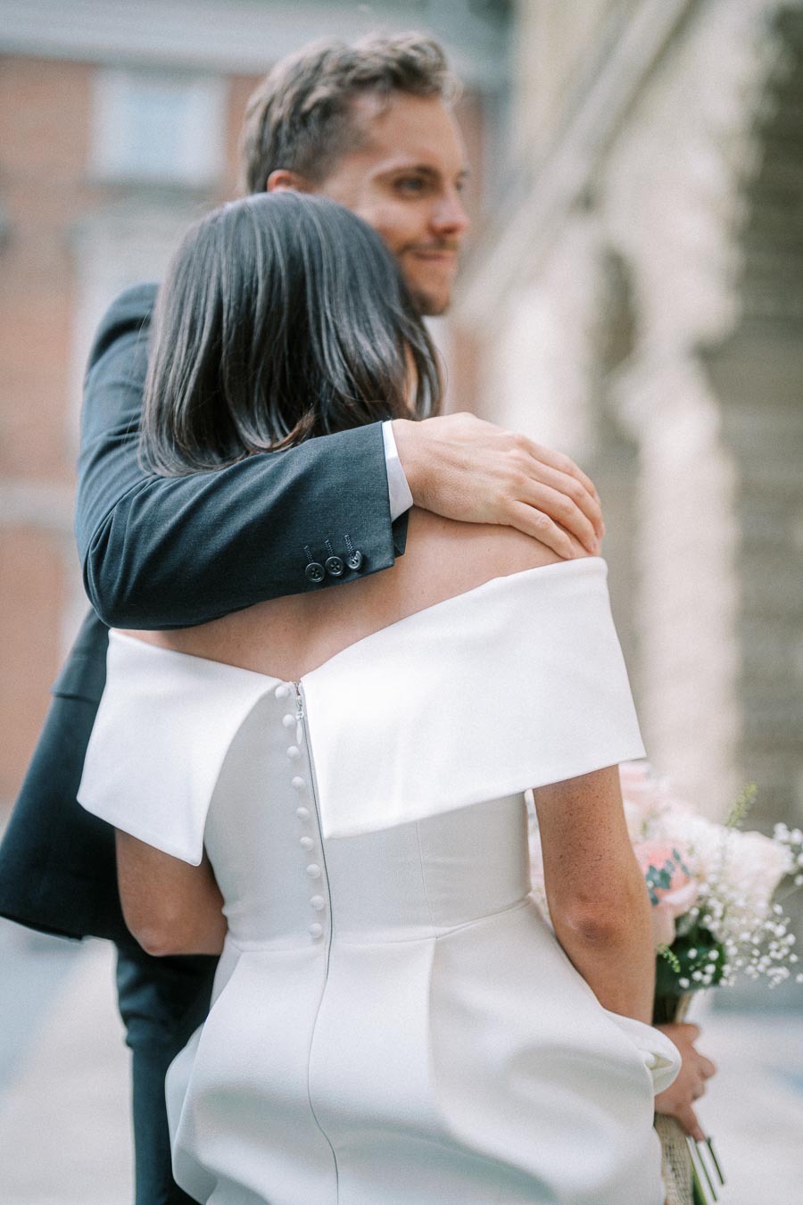 Woman in elegant white wedding dress holding flowers, embraced by man in suit, showcasing romantic outdoor setting.