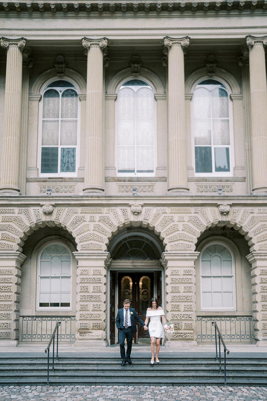 Couple walking down steps of historic building with ornate columns and archways, bride holding bouquet, elegant architectural wedding setting