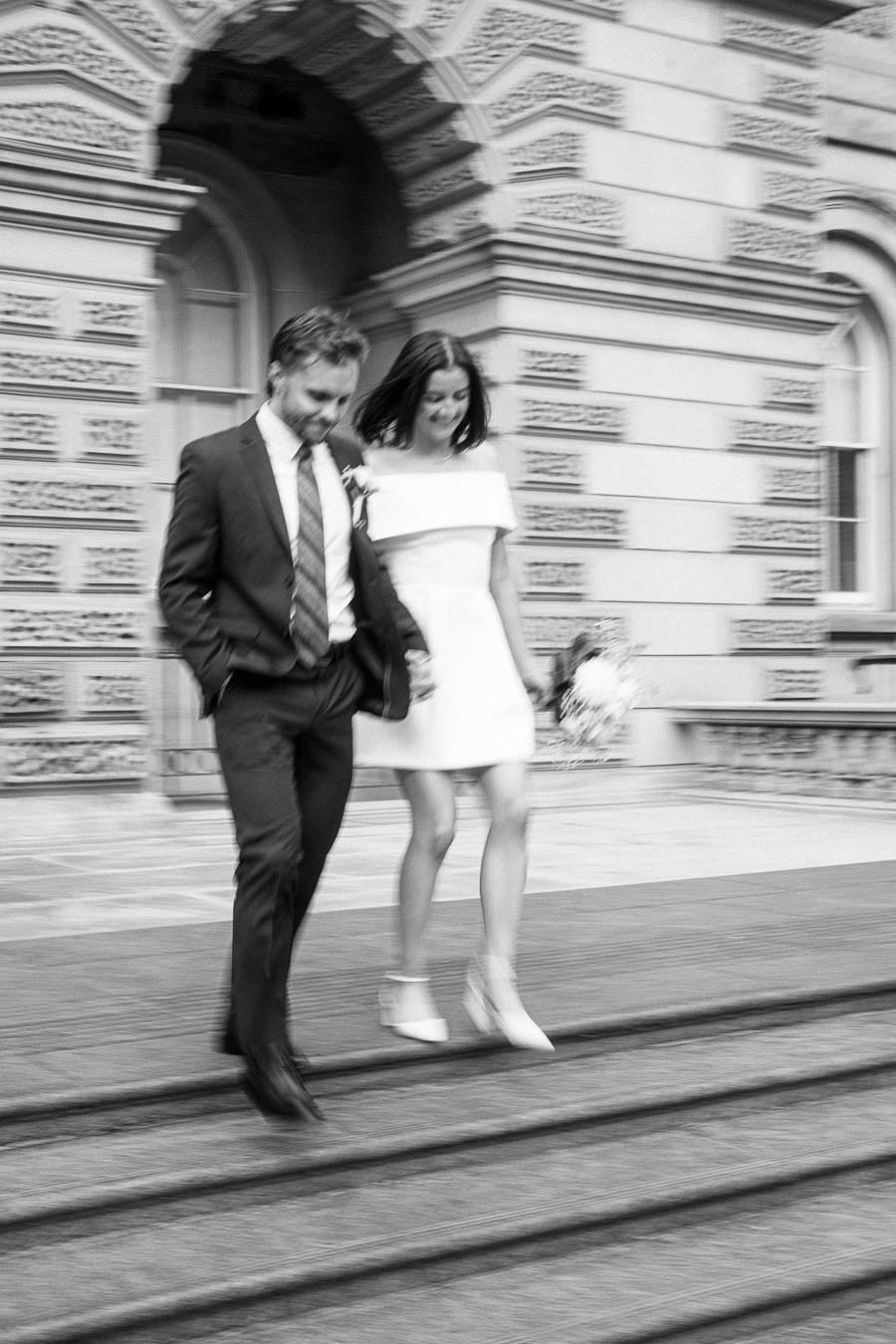 Black and white image of a joyful couple walking down steps, holding hands, outside a historic building with intricate architecture.