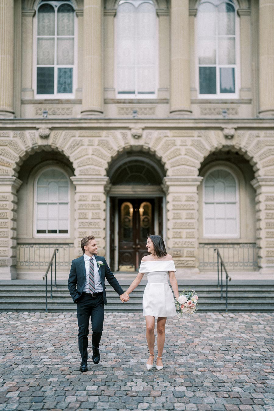 A couple holding hands and smiling in front of a historic building, with the woman wearing a white dress and holding a bouquet.