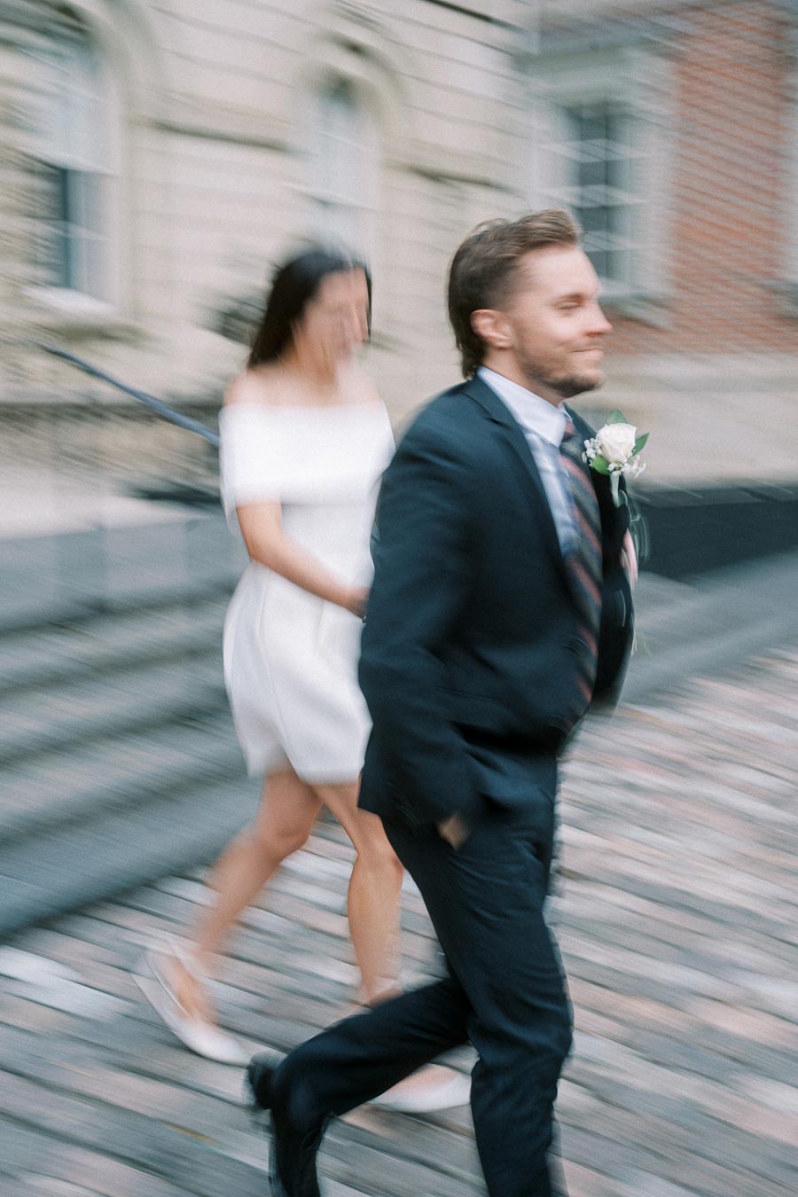 A blurred image of a couple in formal attire walking, with the man in a dark suit and the woman in a white dress, set against an urban backdrop.