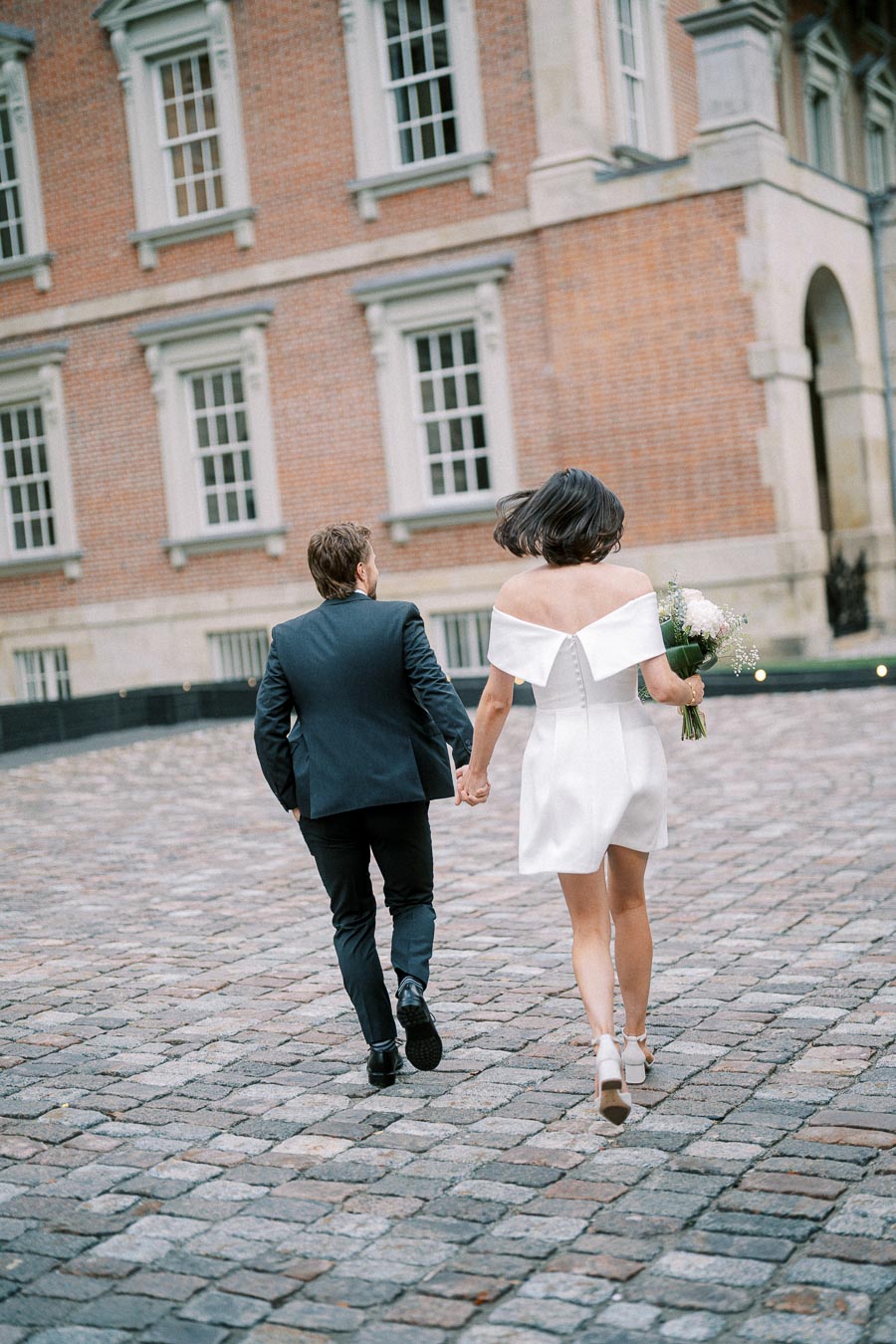 A couple holding hands while walking together on a cobblestone street in front of a historic brick building. The woman is wearing a short white dress holding a bouquet, and the man is in a dark suit.