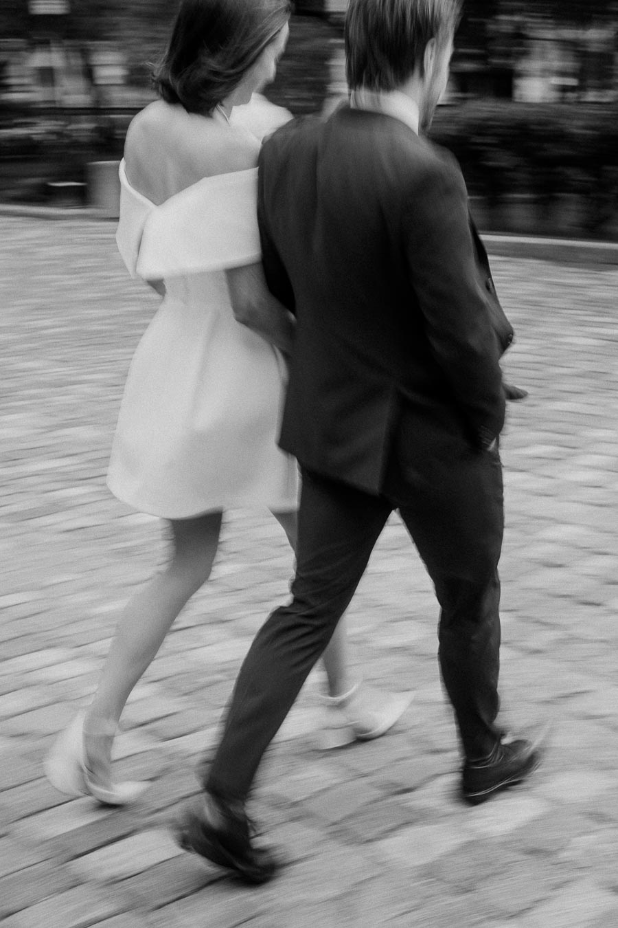 A black and white image of a couple walking together on a cobblestone street. The woman is wearing an elegant off-the-shoulder dress, and the man is in a smart suit. Their movement creates a soft blur, adding a dynamic feel to the photograph.