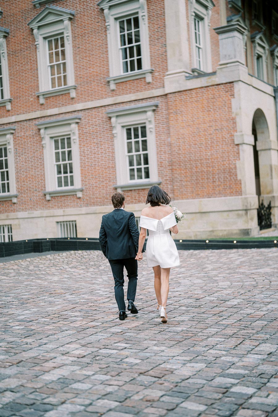 A couple walking hand in hand on a cobblestone path with a historic brick building in the background, dressed in elegant wedding attire.
