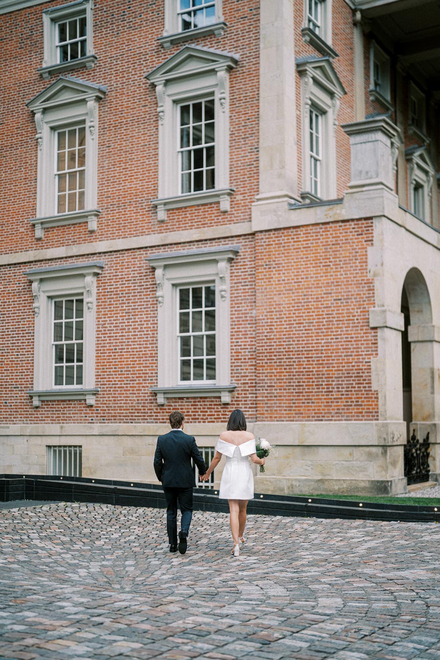 A couple holding hands and walking toward a historic brick building with large windows, the woman in a white dress holding a small bouquet.