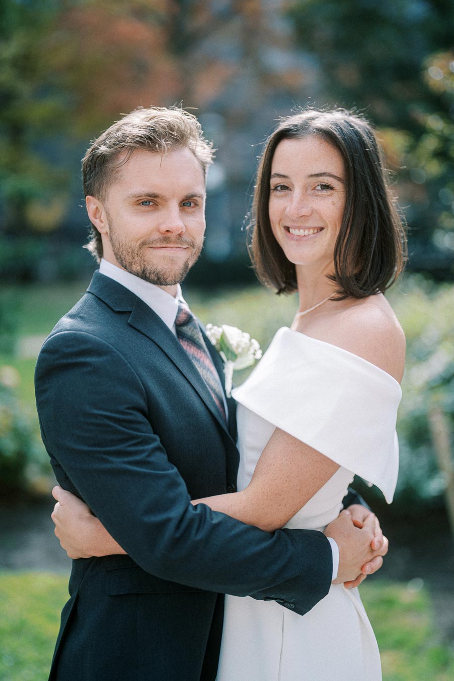 A happy couple embracing in a garden setting, with the man wearing a suit and the woman in an off-shoulder white dress, smiling at the camera.