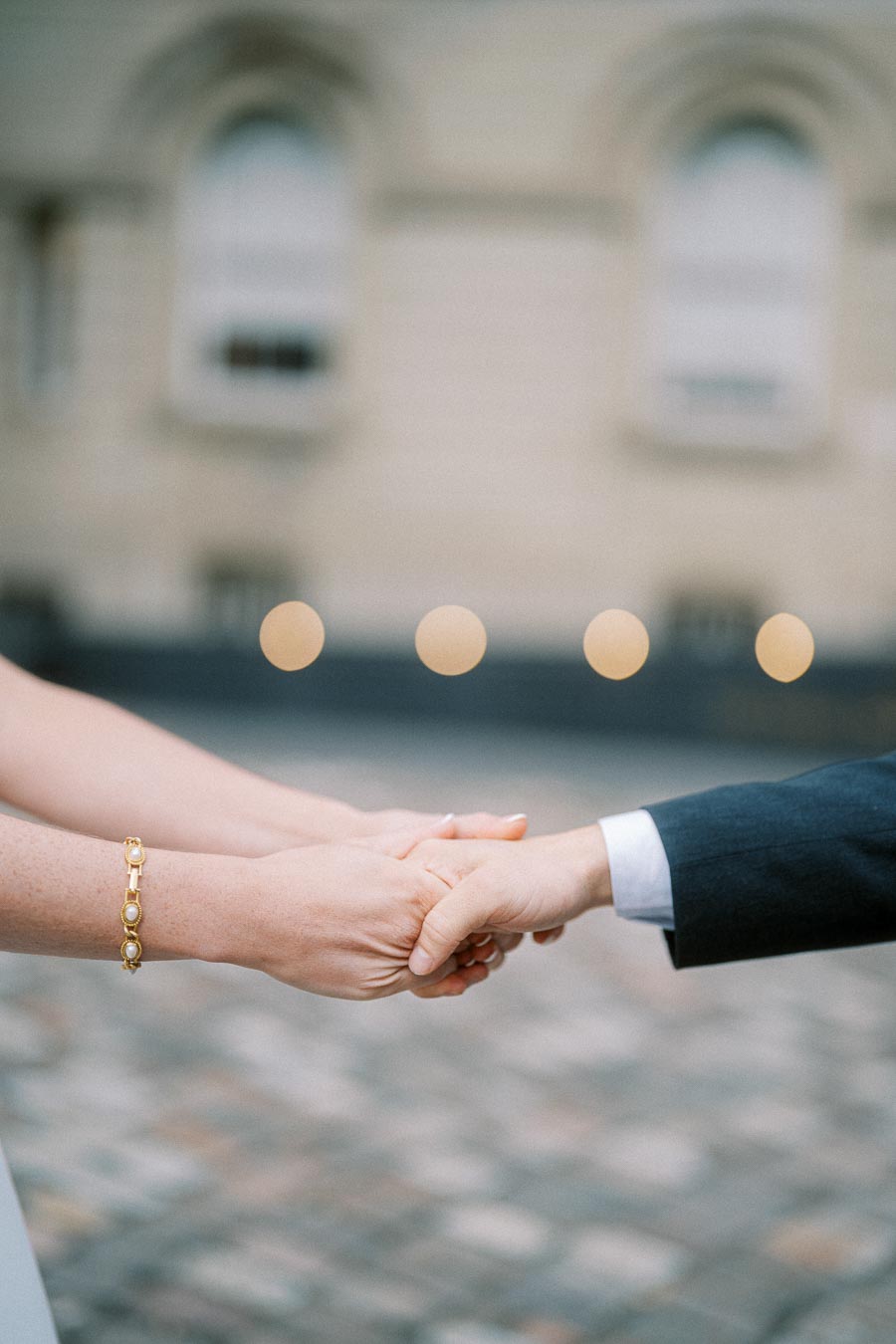 Close-up of a couple holding hands outdoors, featuring a woman wearing a gold bracelet and a man in a dark suit, with a blurred architectural background.