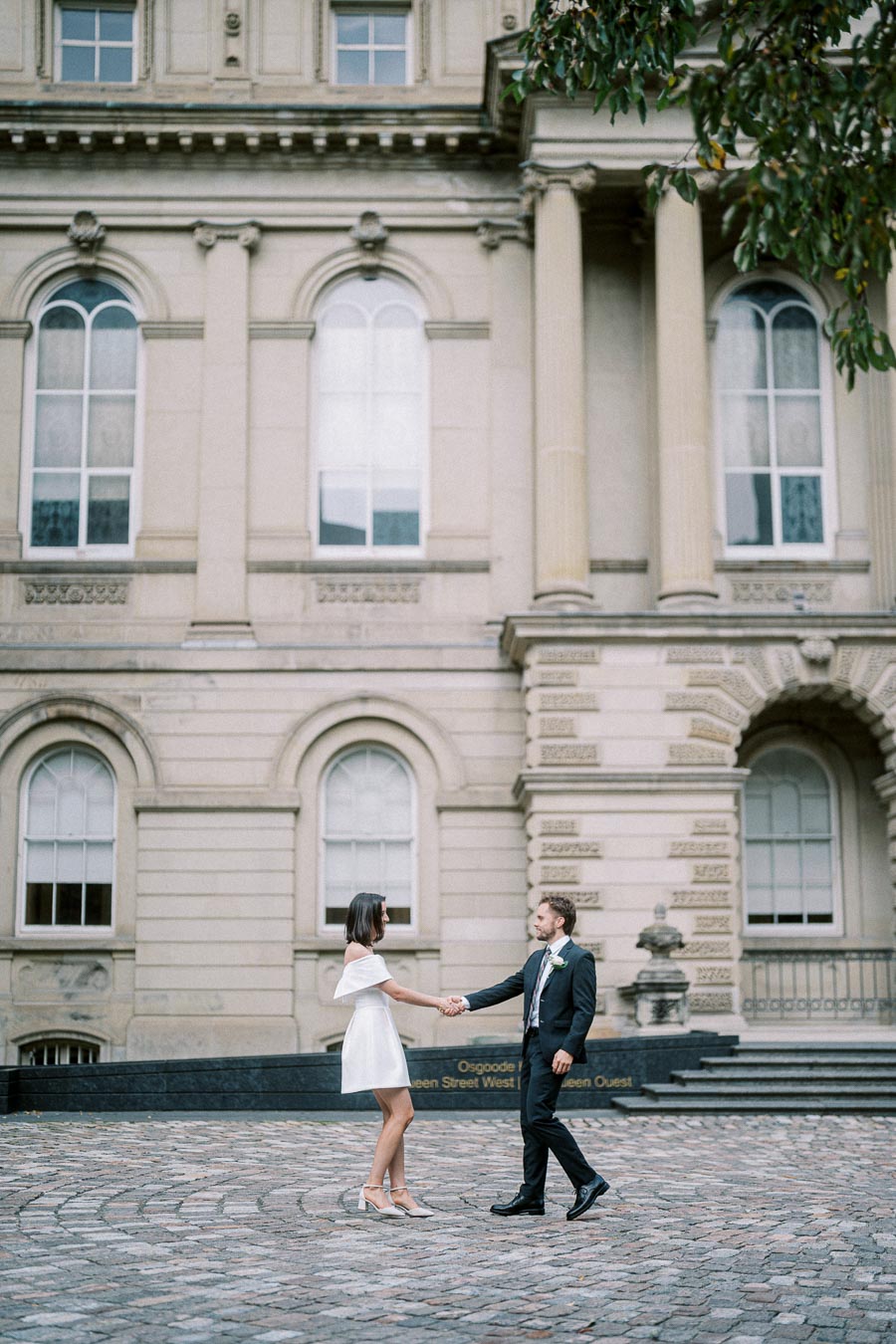 A couple in formal attire holding hands in front of a historic building with arched windows and columns, surrounded by cobblestone pavement.