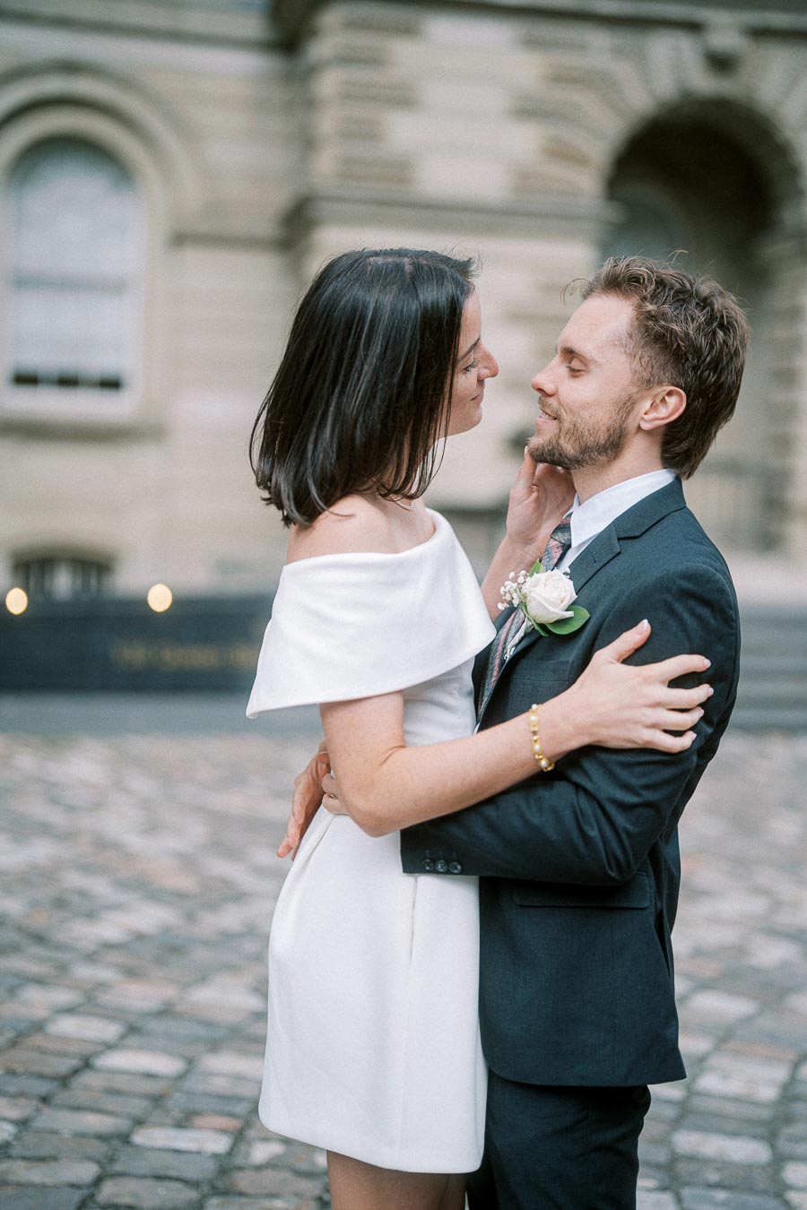 A couple embracing lovingly in front of a historic building with cobblestone pavement, the woman in a white dress and the man in a suit with a boutonniere.