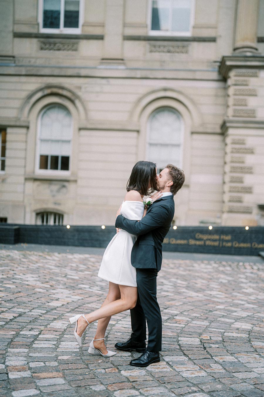 Elegant couple embracing romantically in front of historic building, wearing formal attire with cobblestone pathway underfoot.