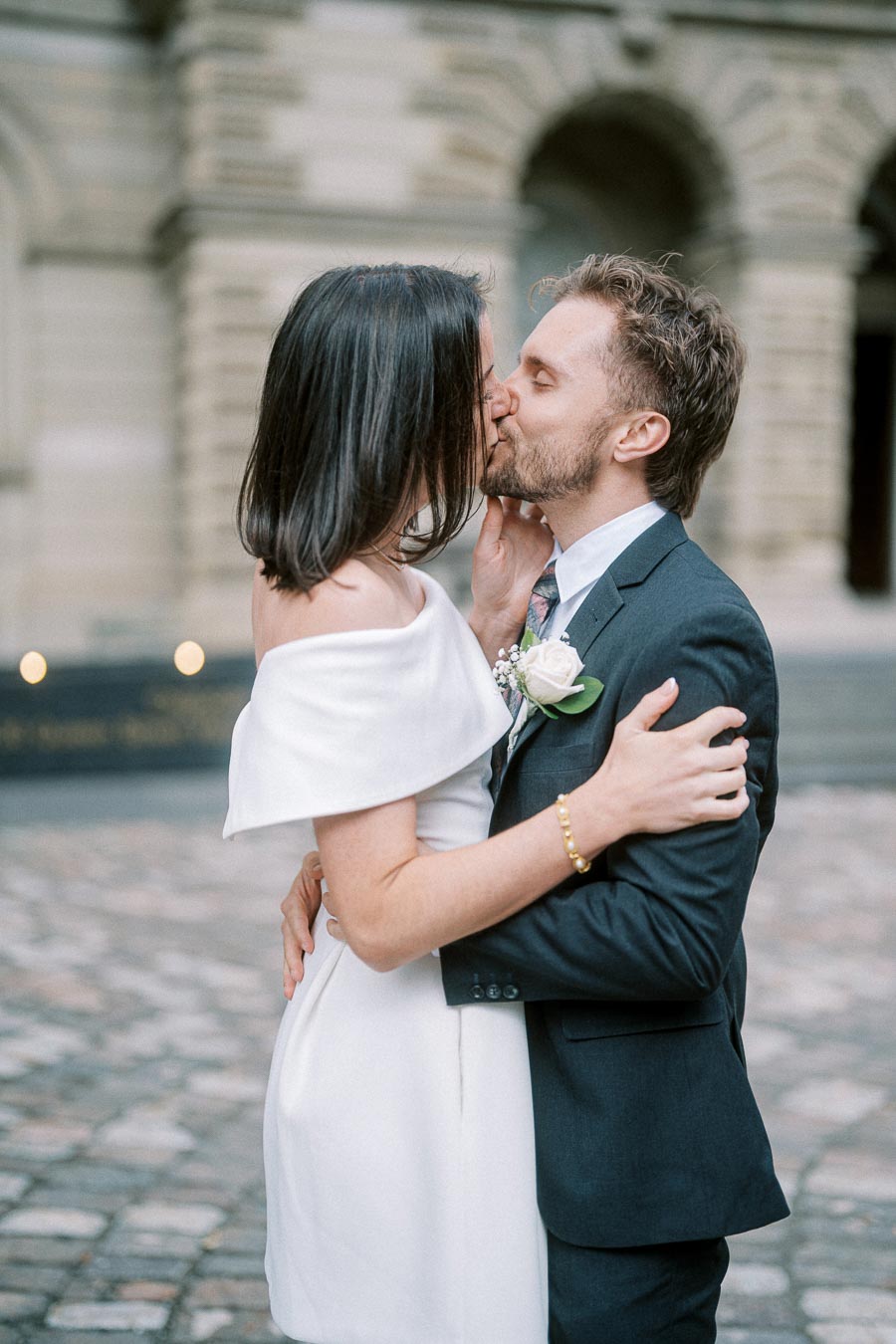 Elegant wedding couple sharing a kiss in front of a historic building, with the bride in an off-shoulder white dress and the groom in a sharp suit, capturing a romantic moment on cobblestone streets