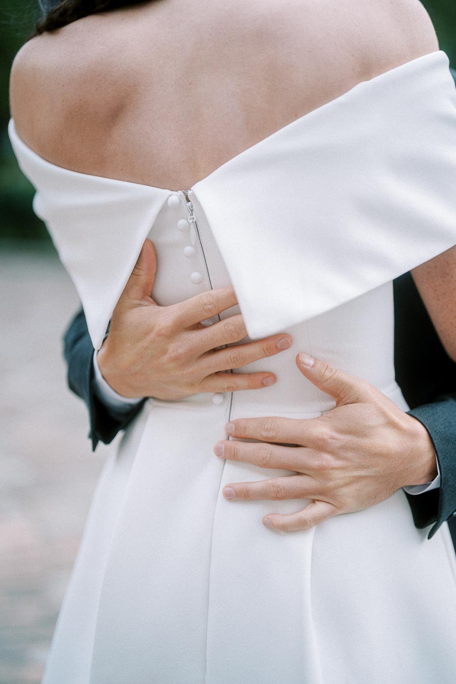 Close-up of a bride and groom embracing, highlighting the back of the bride's elegant off-shoulder wedding dress with button and zip detail.
