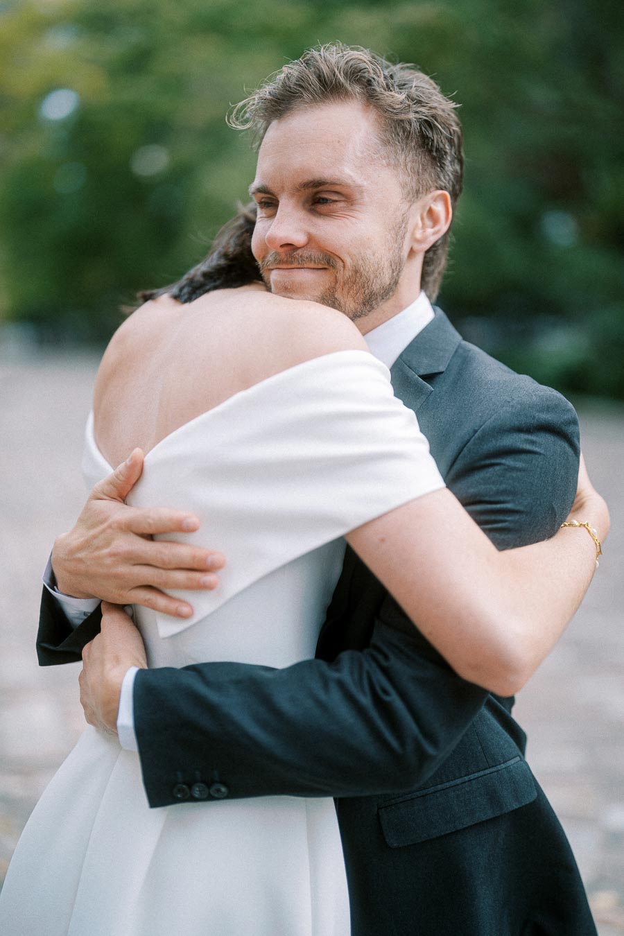 A happy couple embraces outdoors, with the man in a dark suit and the woman in a white off-shoulder dress, surrounded by a natural, blurred background.