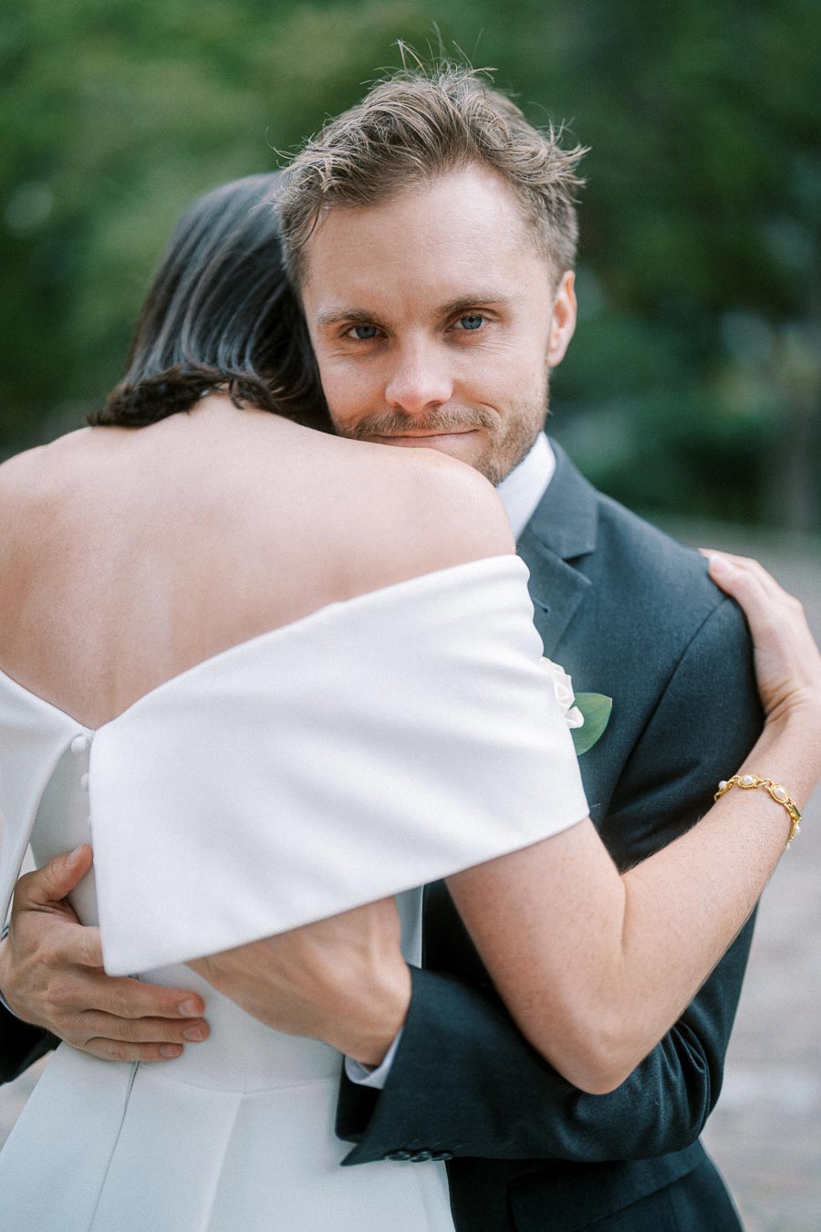 A groom in a dark suit embraces a bride in an off-the-shoulder white dress, looking content and joyful, with a blurred green background.