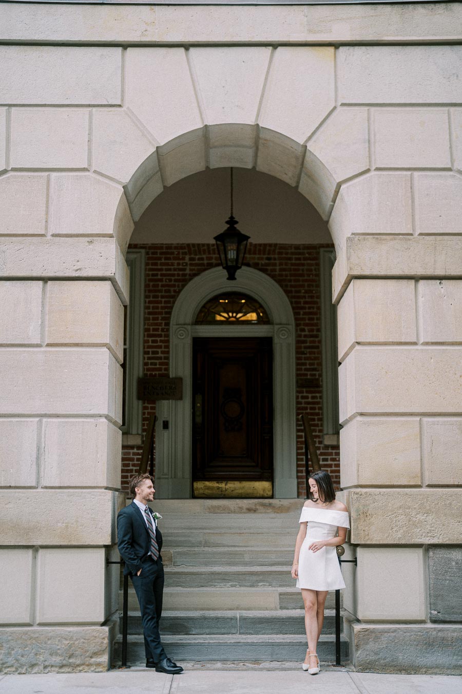 A couple standing under a large stone archway, dressed in formal attire. The man is wearing a dark suit and the woman is in a white dress, smiling at each other. The background features a brick building with an ornate wooden door and hanging lantern.