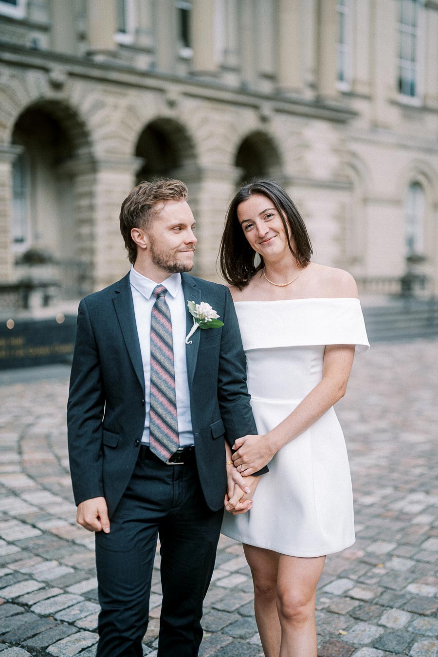 Smiling couple holding hands in elegant wedding attire, posing in front of a historic building with stone arches.