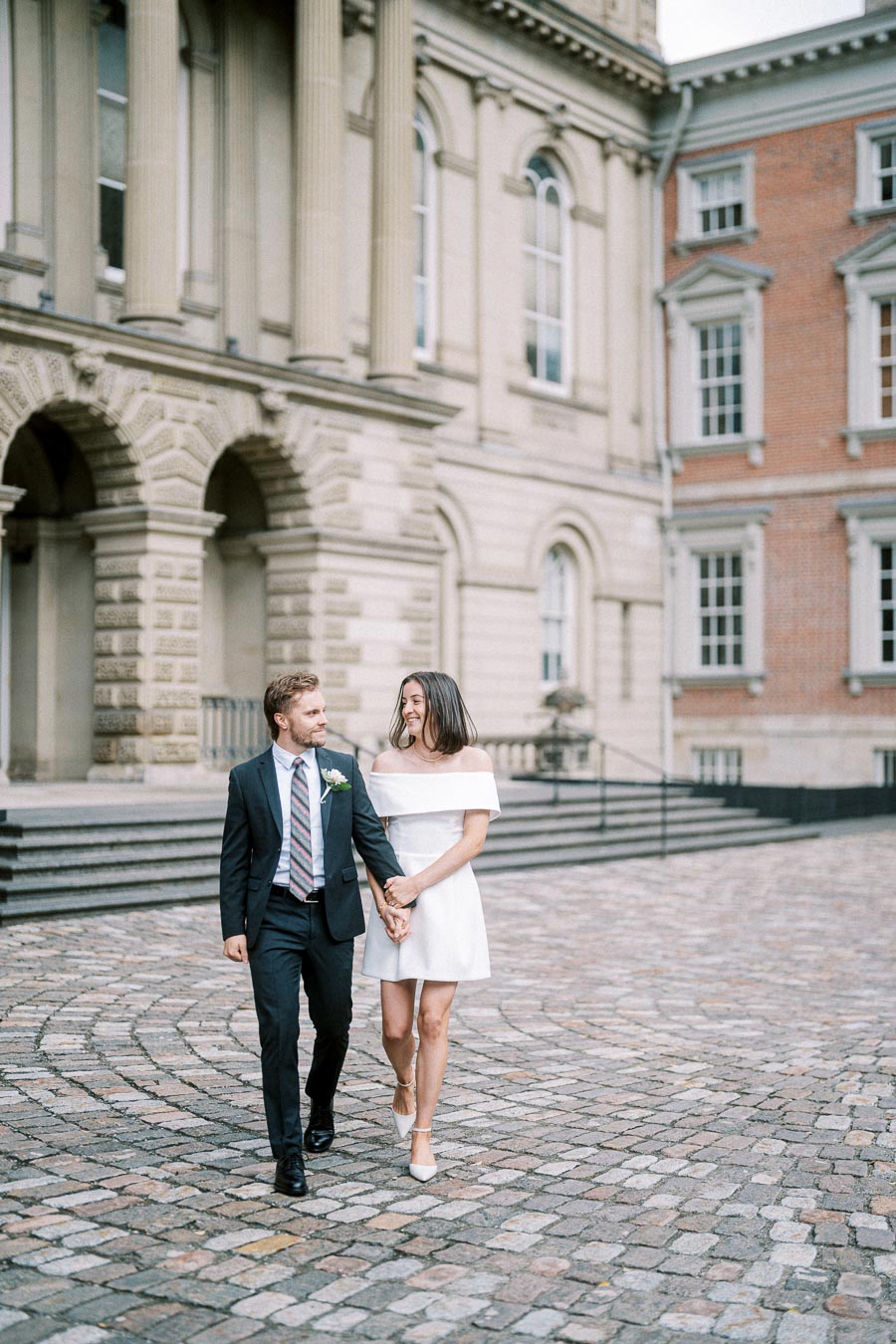 Couple in formal attire walking hand-in-hand outside a historic building with stone architecture.