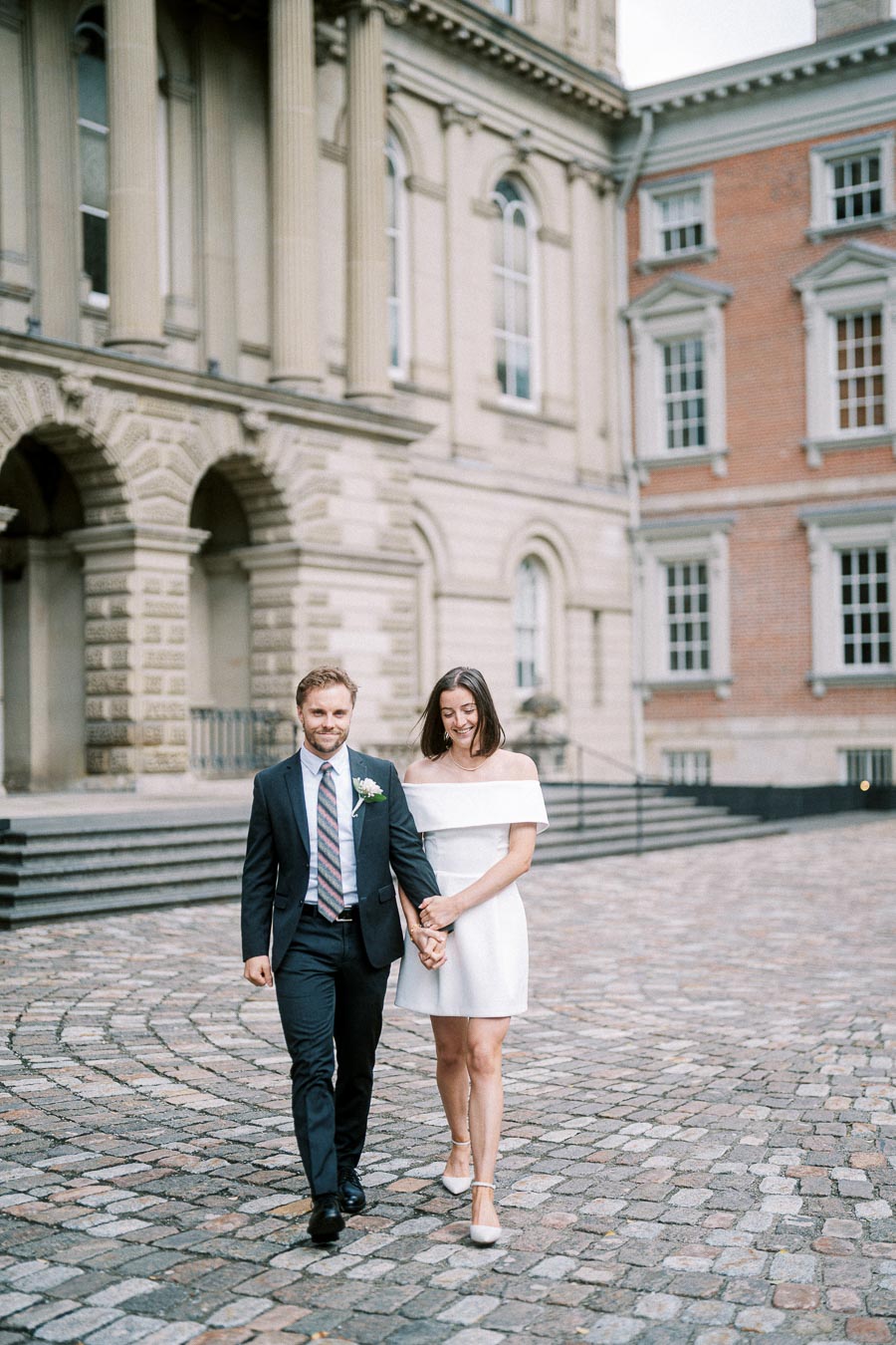 A happy couple in formal attire walking hand in hand on a cobblestone path in front of an elegant historic building.