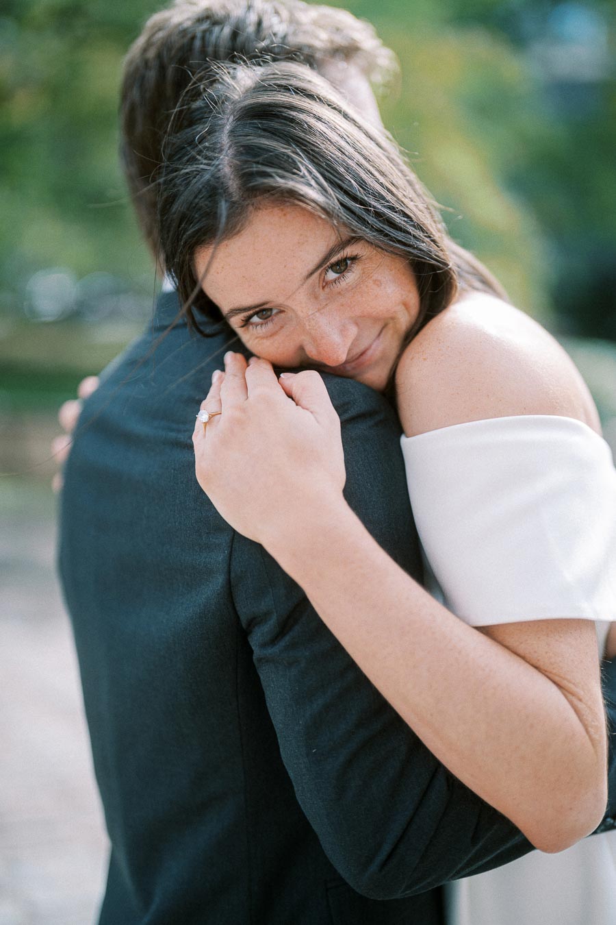 Engaged couple embraces in a tender moment, the woman wears an off-shoulder white dress and shows her engagement ring while smiling softly, surrounded by a blurred natural background.