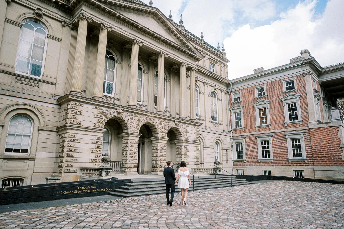 Osgoode Hall entrance with a couple walking towards the historic architecture, showcasing grand columns and detailed brickwork on a sunny day in Toronto.
