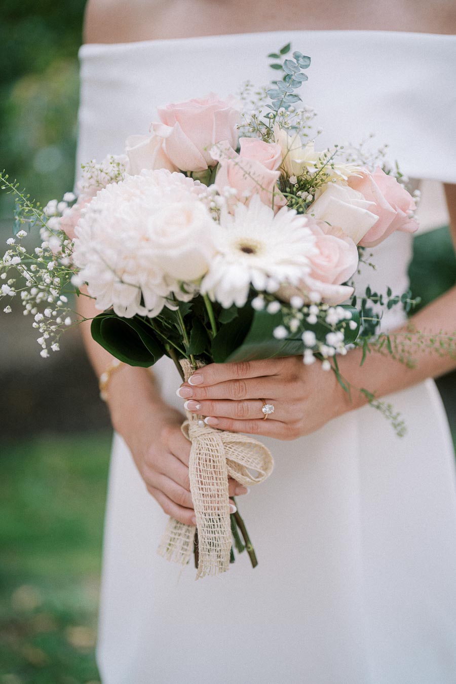 A bride in a white dress holds a bouquet of soft pink roses, white daisies, and greenery, tied with a burlap ribbon, showcasing an elegant wedding arrangement.