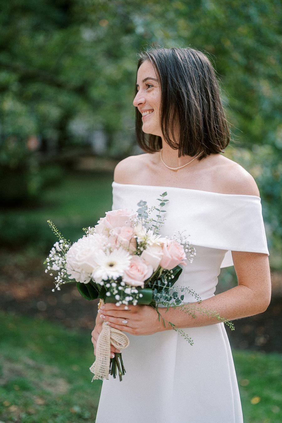 A smiling bride in an elegant off-shoulder wedding dress holds a bouquet of pale pink and white flowers, standing in a lush green garden.