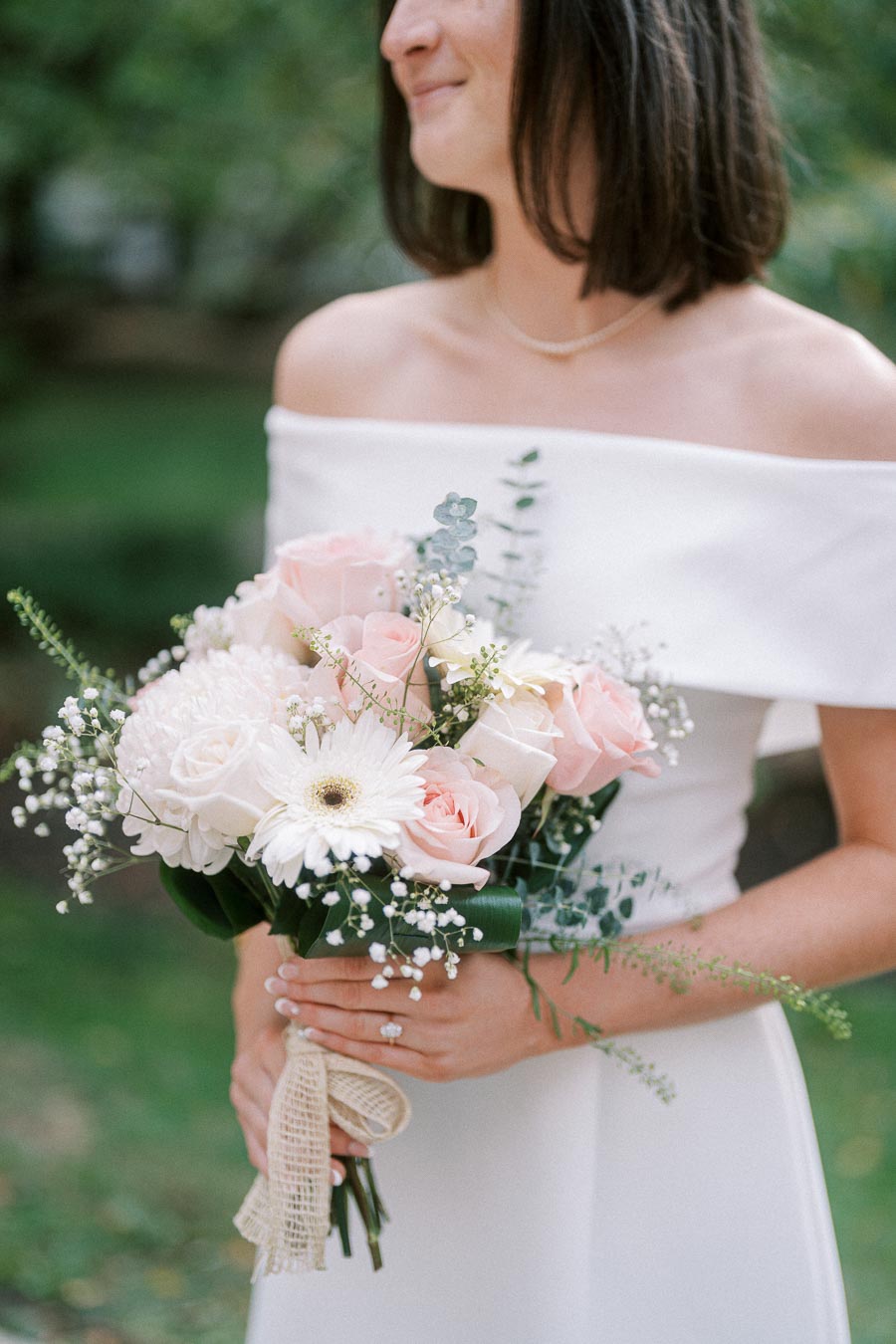 A bride in a white dress holding a bouquet of pink and white flowers with greenery, outdoors in a garden setting.