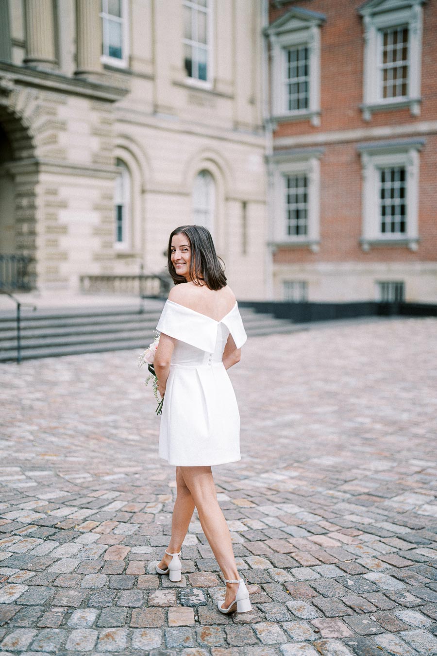 Young woman in a white dress smiling while walking on cobblestone street in front of a historic building, holding flowers.