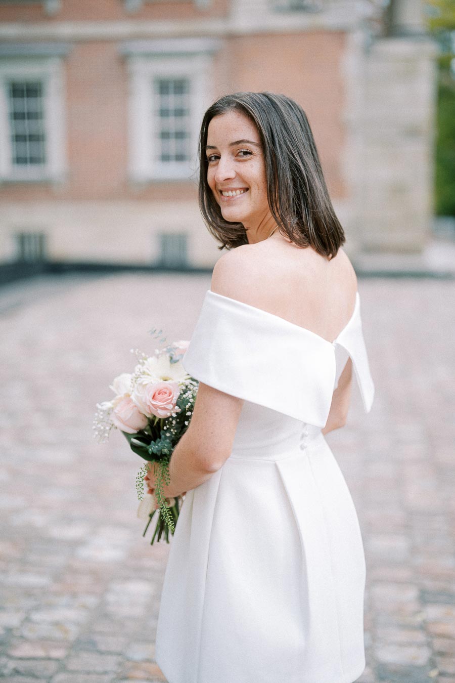 Smiling bride in an elegant off-shoulder white wedding dress holding a bouquet of pink and white roses, standing on a cobblestone pathway in front of a historic building.