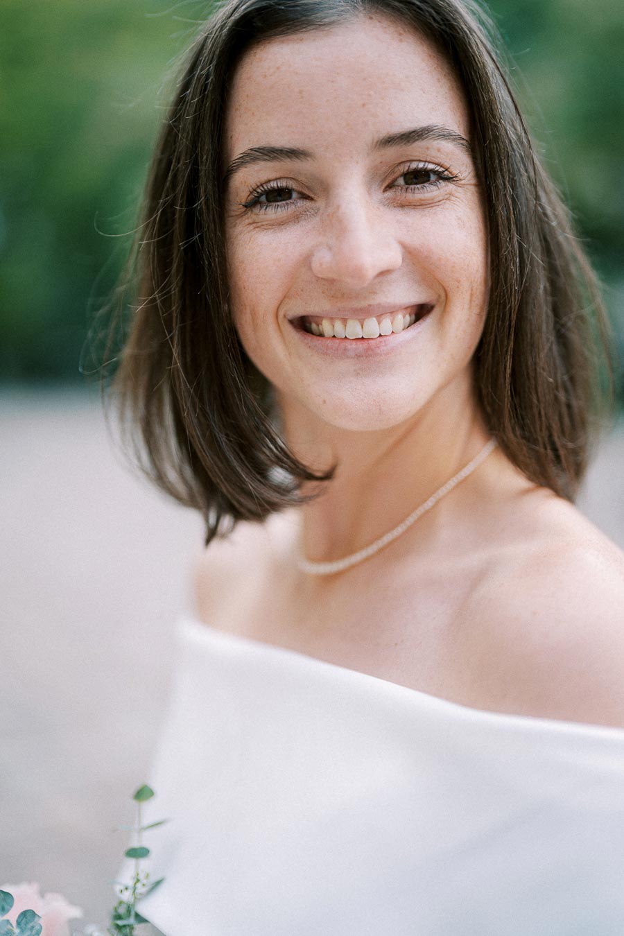 A smiling woman with shoulder-length brown hair and a white top standing outside with greenery in the background.