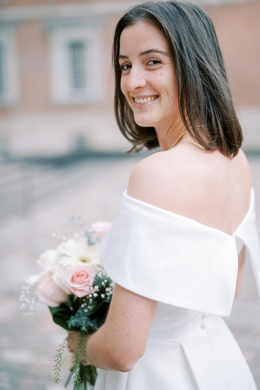 A bride in an elegant off-shoulder white wedding dress smiles while holding a bouquet of pastel pink and white flowers, with a blurred building background.