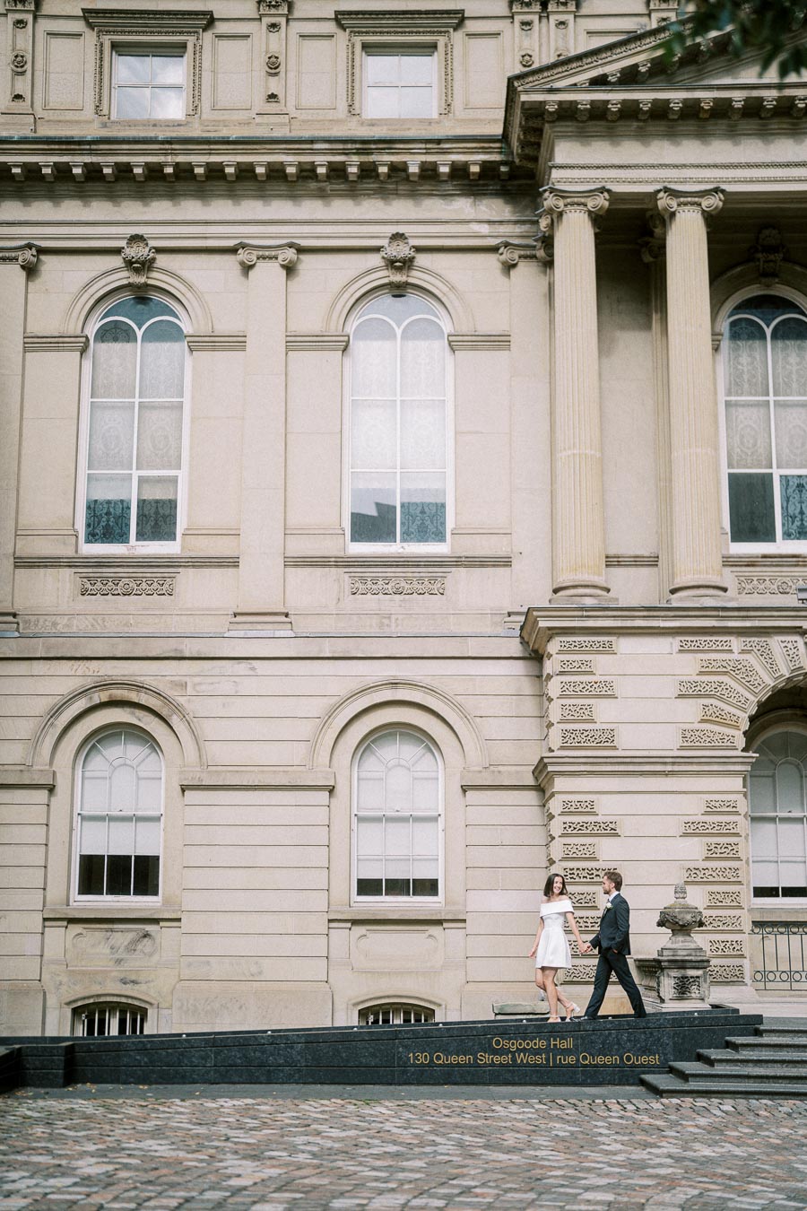 Couple walking in front of historic Osgoode Hall on Queen Street West, showcasing architectural details and romantic ambiance
