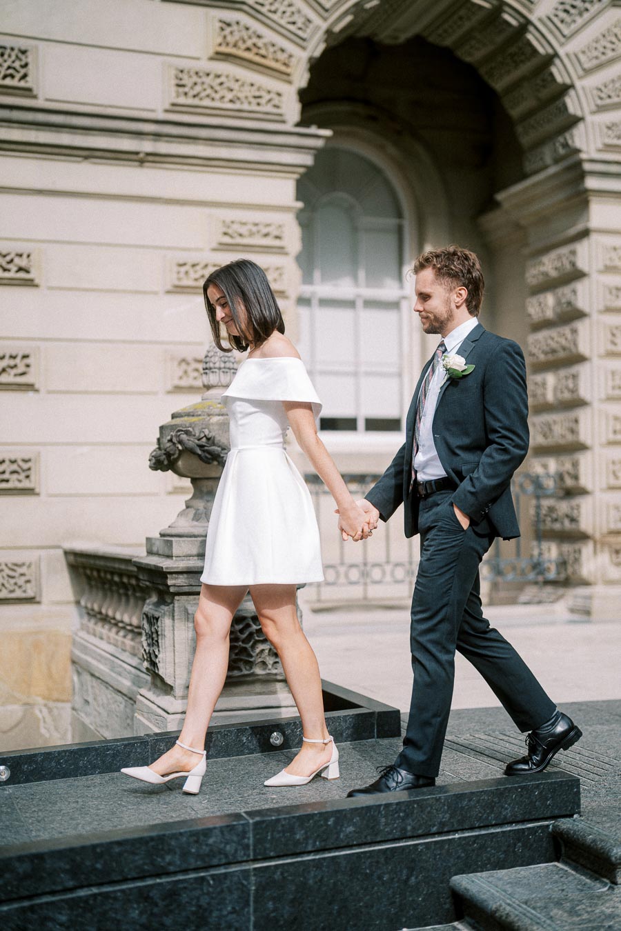 A couple holding hands and walking down stone steps, with the woman in a white dress and the man in a suit with a boutonnière, in front of an ornate historical building.