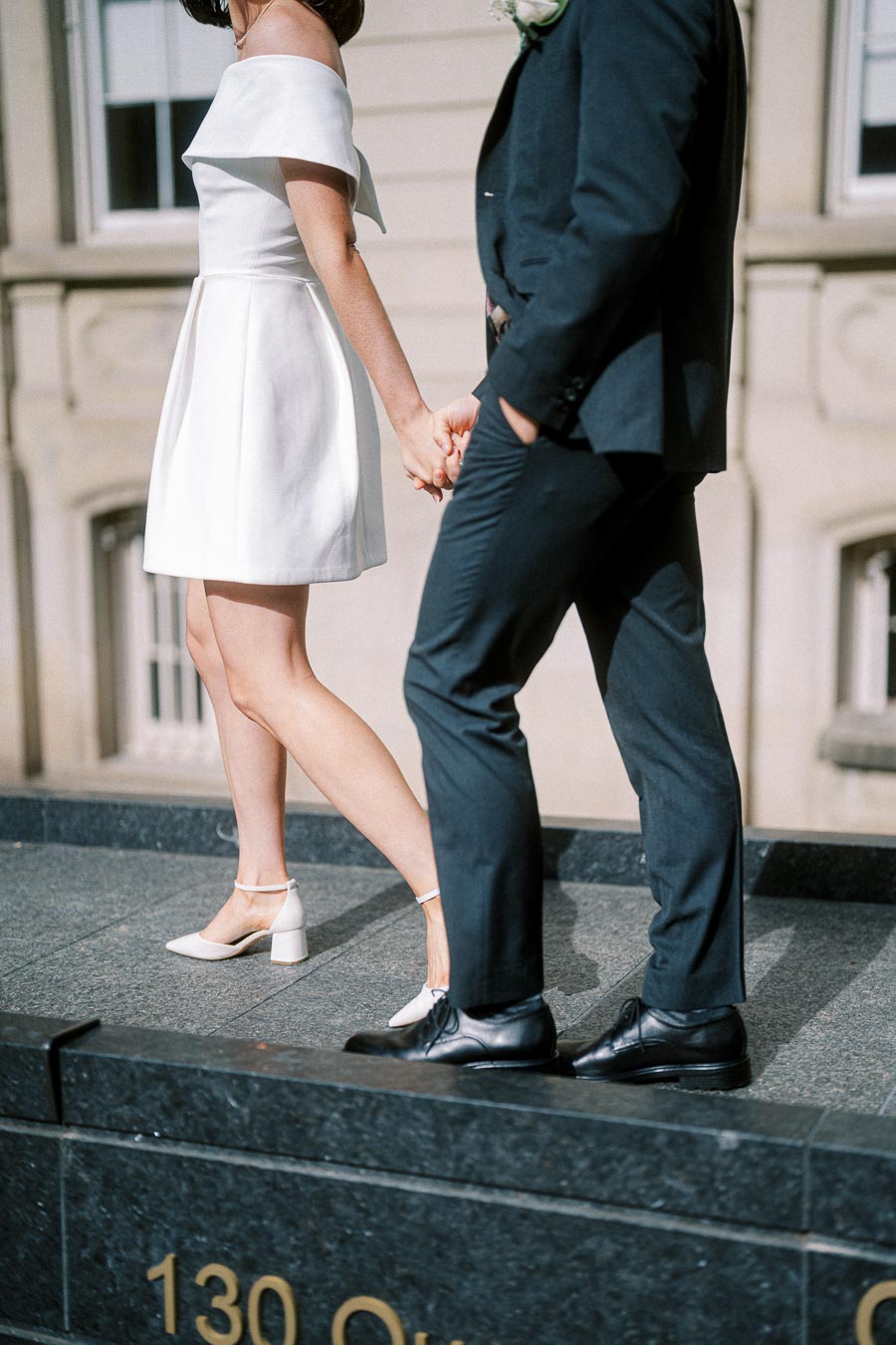 A stylish couple holds hands while walking on a city street, with the woman wearing an elegant white dress and the man in a classic black suit.