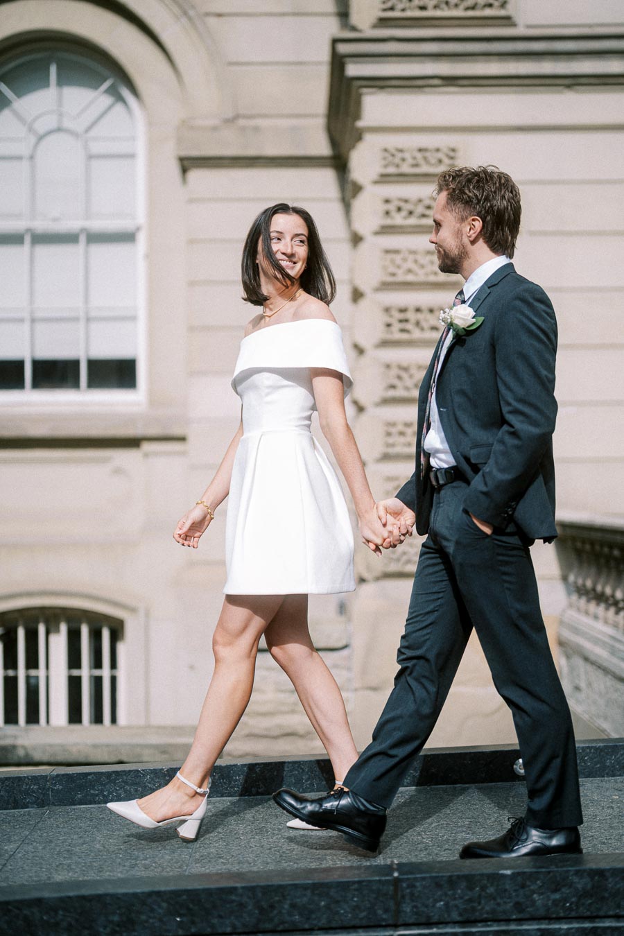 A couple walking hand in hand in formal attire, with the woman in a white dress and the man in a dark suit, set against an elegant, historic building background.