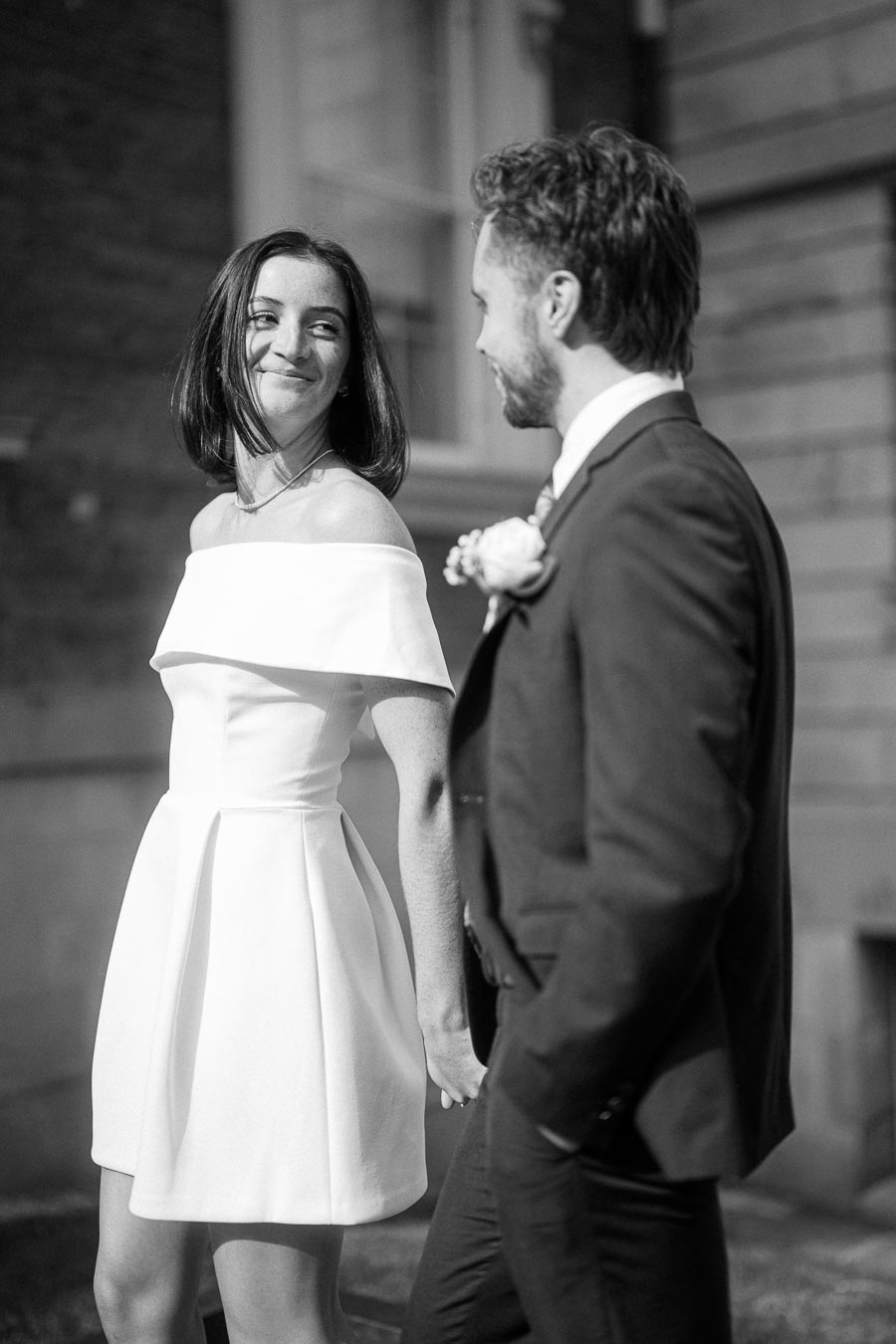 Black and white photo of a smiling couple holding hands, with the woman in a white off-shoulder dress and the man in a suit, captured outdoors.
