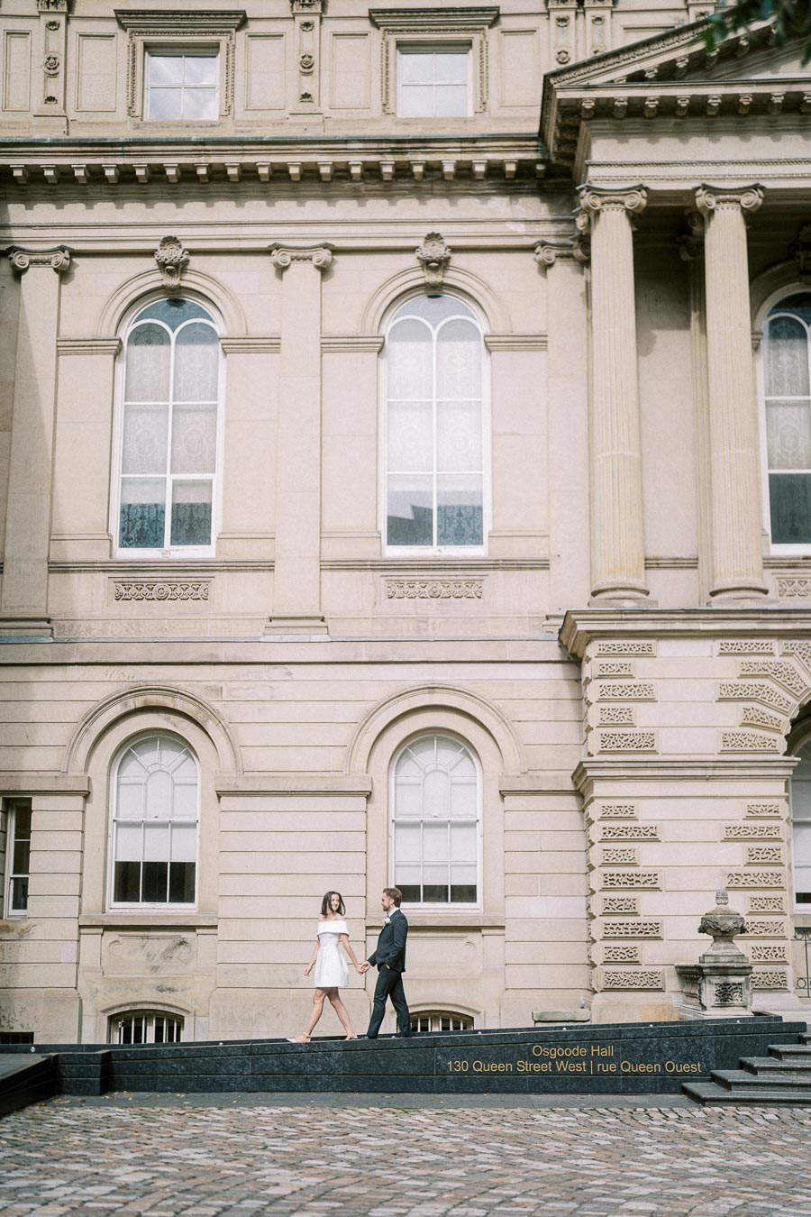 A couple walking hand in hand in front of the historic architecture of Osgoode Hall, 130 Queen Street West, showcasing detailed stonework and arched windows.