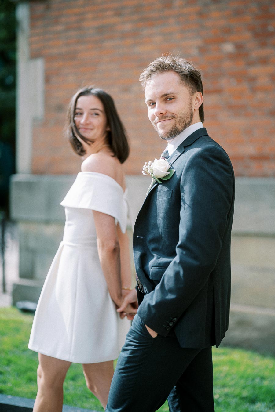 Couple smiling and holding hands during a wedding photo shoot, with the groom in a dark suit and boutonniere, and the bride in an off-shoulder white dress, standing in front of a brick wall.