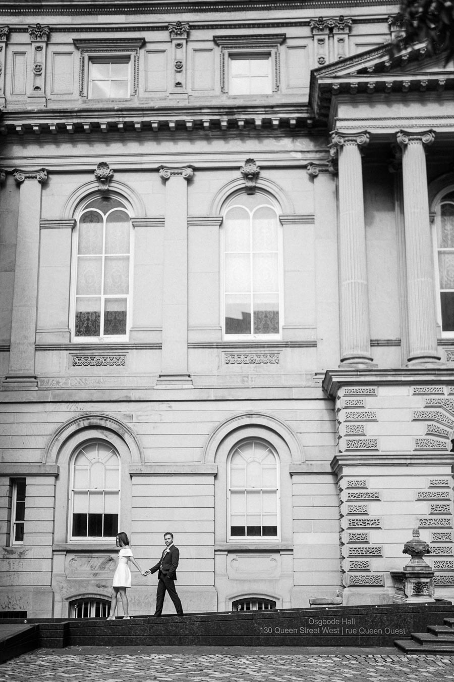 Black and white photo of a couple holding hands and walking in front of the historic Osgoode Hall building, featuring large windows and detailed architectural elements.