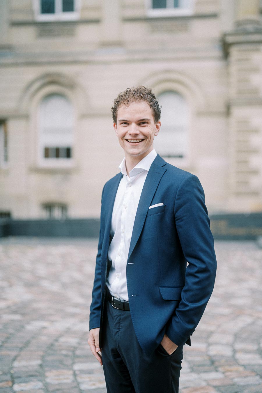 A smiling man in a blue suit stands outdoors in front of a historical building with stone architecture.