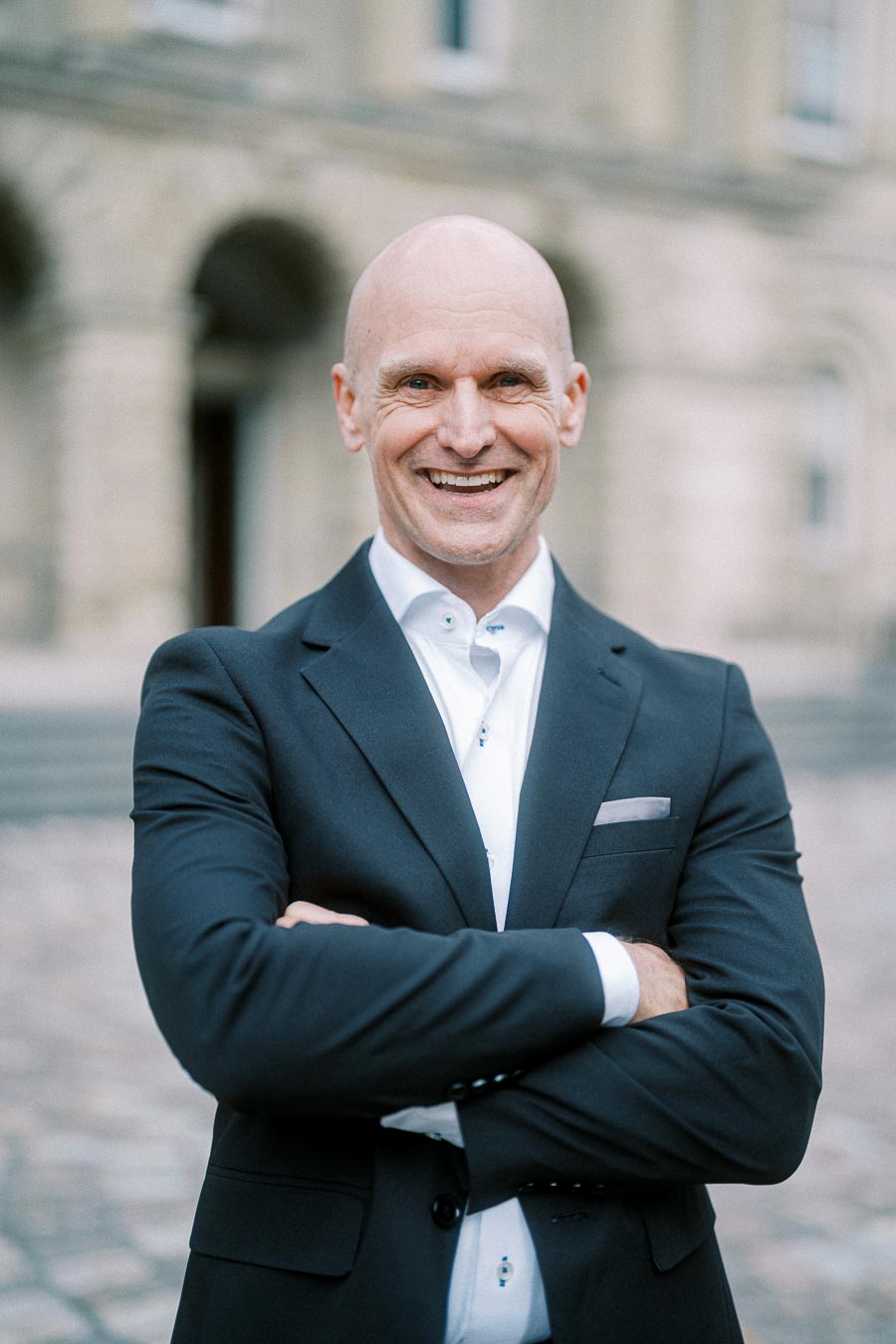 Smiling man in a dark suit with arms crossed, standing in front of a historical building.